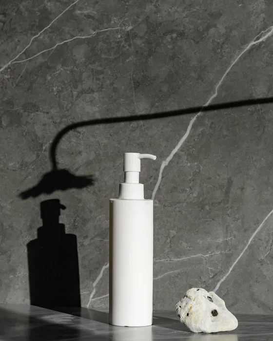A white pump bottle on dark marble, lit to throw a hard, sculpted shadow.