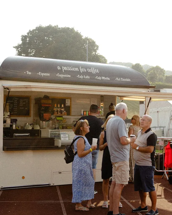 A white catering trailer parked on grass with a queue of people waiting at the serving hatch.